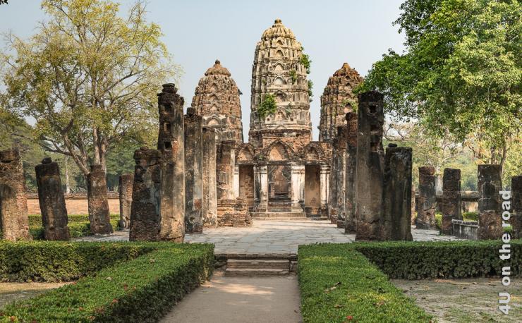 Bild Tempel im Historical Park - Sukhothai zeigt im Hintergrund den Tempel mit drei Türmen. Der Weg hin ist gesäumt von niedrigen Hecken und hohen Säulen. Rechts und links stehen niedrigere Säulen und Bäume.