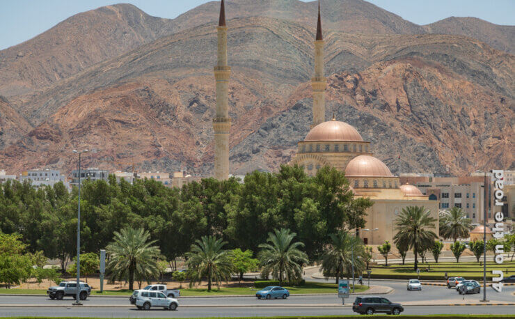 Die spektakuläre Moschee passt sich farblich perfekt dem Felsen im Hintergrund an. Im Vordergrund befinden sich gepflegte Rasenflächen, Palmen und schattenspendende Bäume.