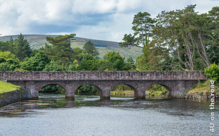 Eine alte Steinbrücke mit vier Bögen überquert den Fluss. Dahinter wachsen urige Bäume und ragen kahle Hügel empor - Autofahren in Irland