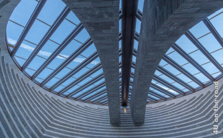 Das Bild zeit das runde Dach der Kirche Mogno. Der blaue Himmel wird von einem Kondensstreifen durchzogen. Die Stützpfeiler, auch als Himmelsleiter zu sehen wölben sich unter dem schrägen Dach aus Glas und Stahl.